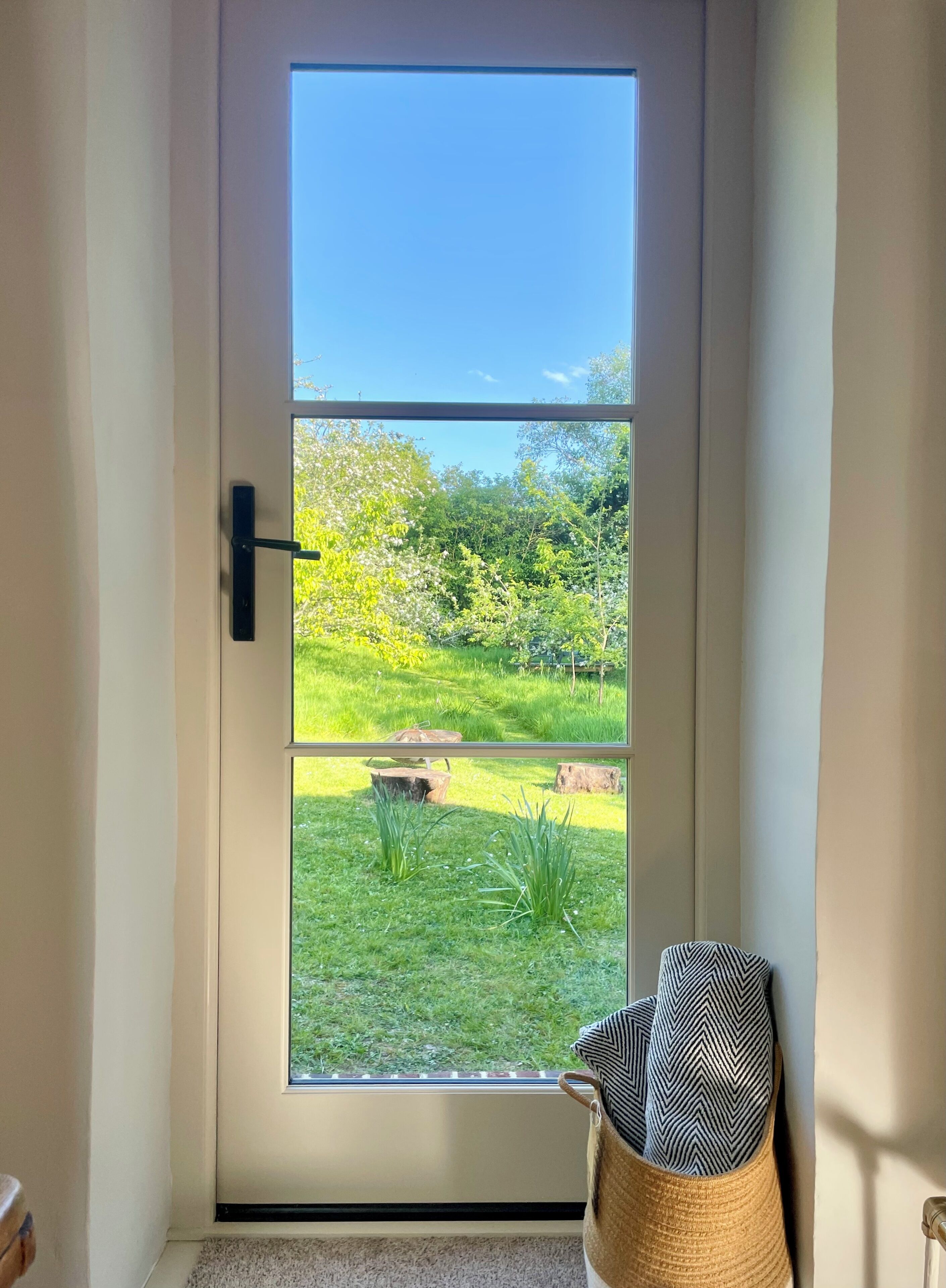 A door with a view of a garden, a black handle, and a woven basket with a patterned pillow.