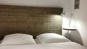 A bed with white linens, a wicker headboard, and a bedside table with a lamp. - Ferienwohnung Schuppen XII mit Terrasse (Norderney)