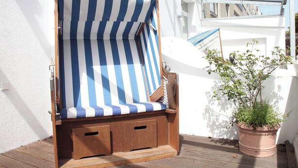 A wooden beach chair with a blue and white striped canopy, positioned on a wooden deck next to a white wall and a potted plant. - Ferienwohnung Schuppen XII mit Terrasse (Norderney)