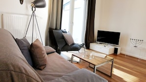 A modern living room with a sofa, a coffee table, a TV, and a floor lamp. - Ferienwohnung Schuppen XII mit Terrasse (Norderney)