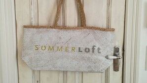 A white door with a woven-textured bag hanging on it, labeled "SOMMERLOFT". - Ferienwohnung Schuppen XII mit Terrasse (Norderney)