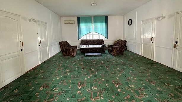 A room with a patterned carpet, a bed with a headboard, two chairs, a window with curtains, and a clock on the wall. - Sunny Hostel (Osh)