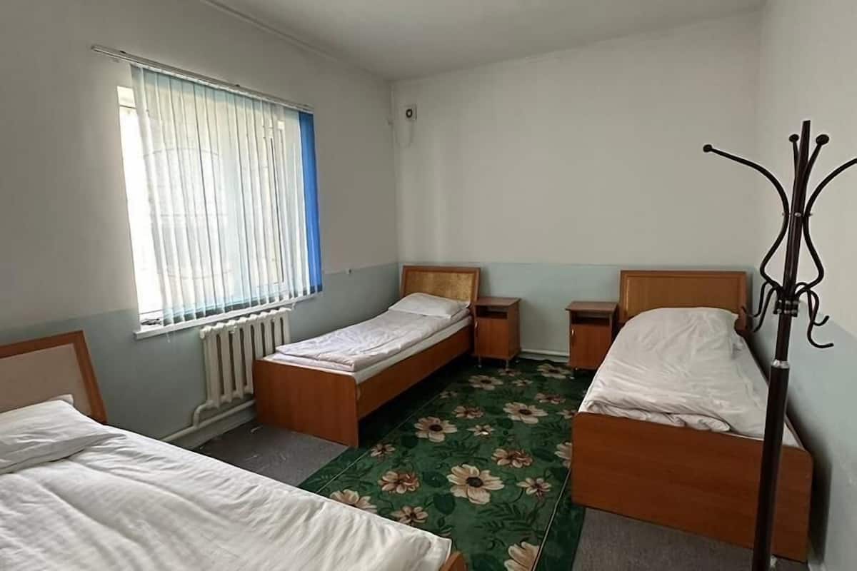 Habitación con tres camas individuales, un radiador, una ventana con persianas y un perchero, además de una alfombra verde con estampado floral.