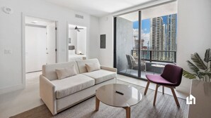 A modern living room with a sofa, a coffee table, and a chair. There is a large window with a view of high-rise buildings. - High-Rise Balcony: Ocean Views, Pool & Spa (Miami)