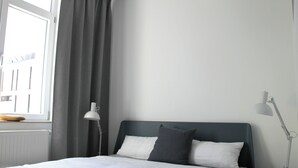 Cama bem arrumada com roupa de cama branca e cinza, cabeceira cinza e mesa de cabeceira com abajur.