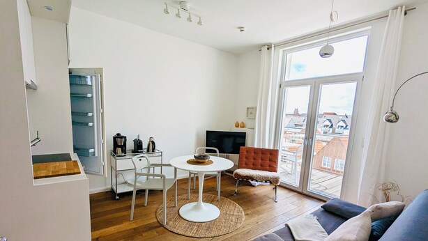 Uma sala de estar moderna com sofá, mesa de centro e televisão. Há uma varanda com vista para edifícios.