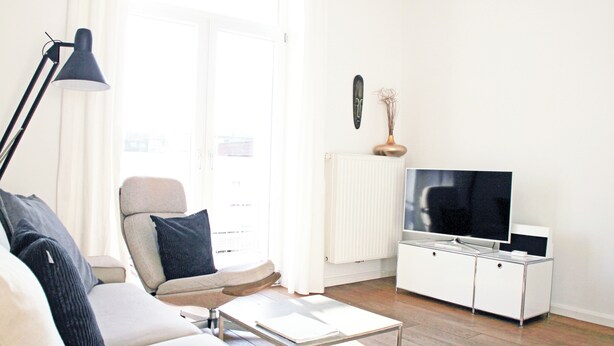 Een moderne woonkamer met een bank, een salontafel, een televisie op een wit kastje en een vloerlamp.