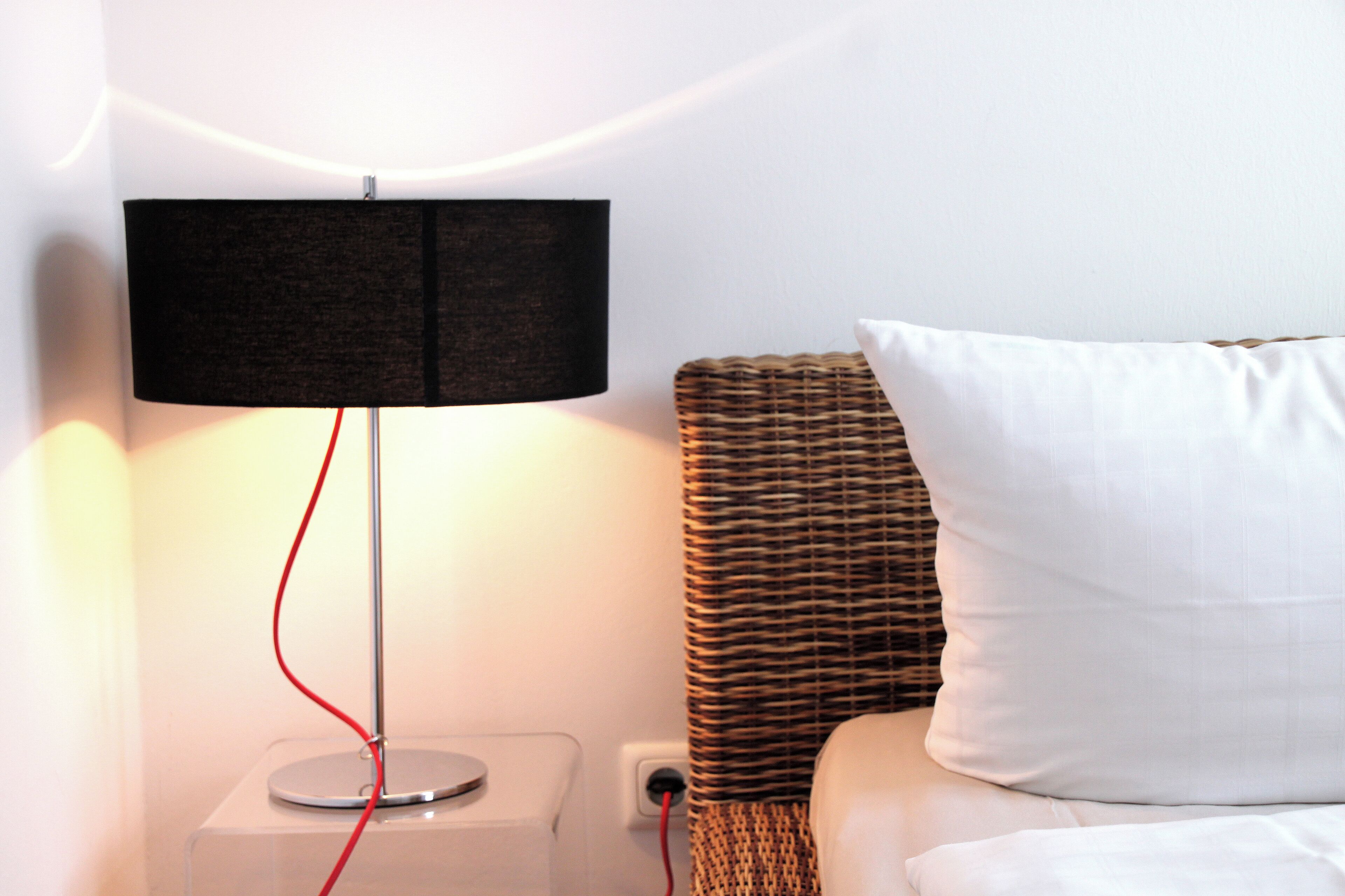 A modern room with a clear nightstand, a lamp with a red cord, a white pillow, and a wicker headboard.