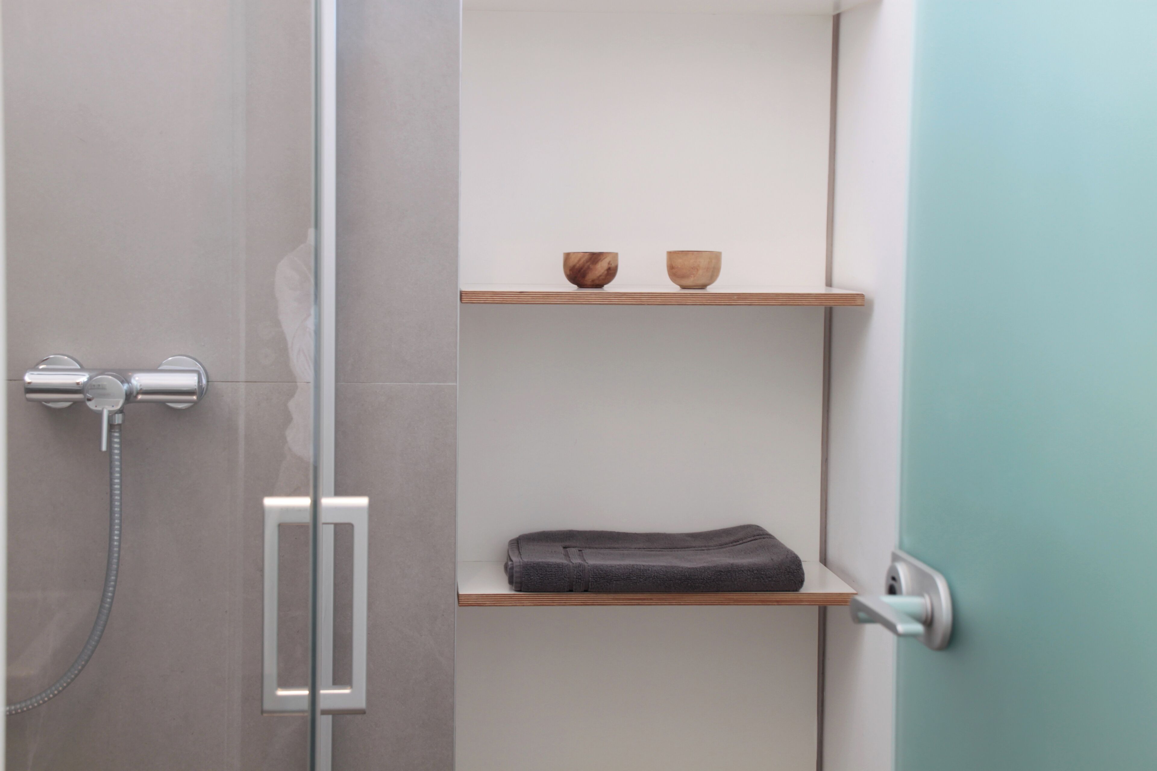 A bathroom with a glass shower enclosure, a towel rack, and two wooden shelves with bowls and a folded towel.