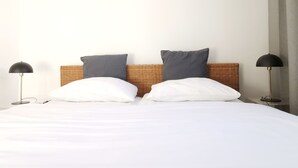 Una cama bien tendida con sábanas blancas y dos almohadas, un cabecero de mimbre y dos lámparas de pie negras a cada lado.