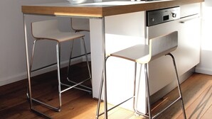 Una cocina compacta con encimera de madera, dos taburetes blancos tipo bar, un microondas y una cafetera.