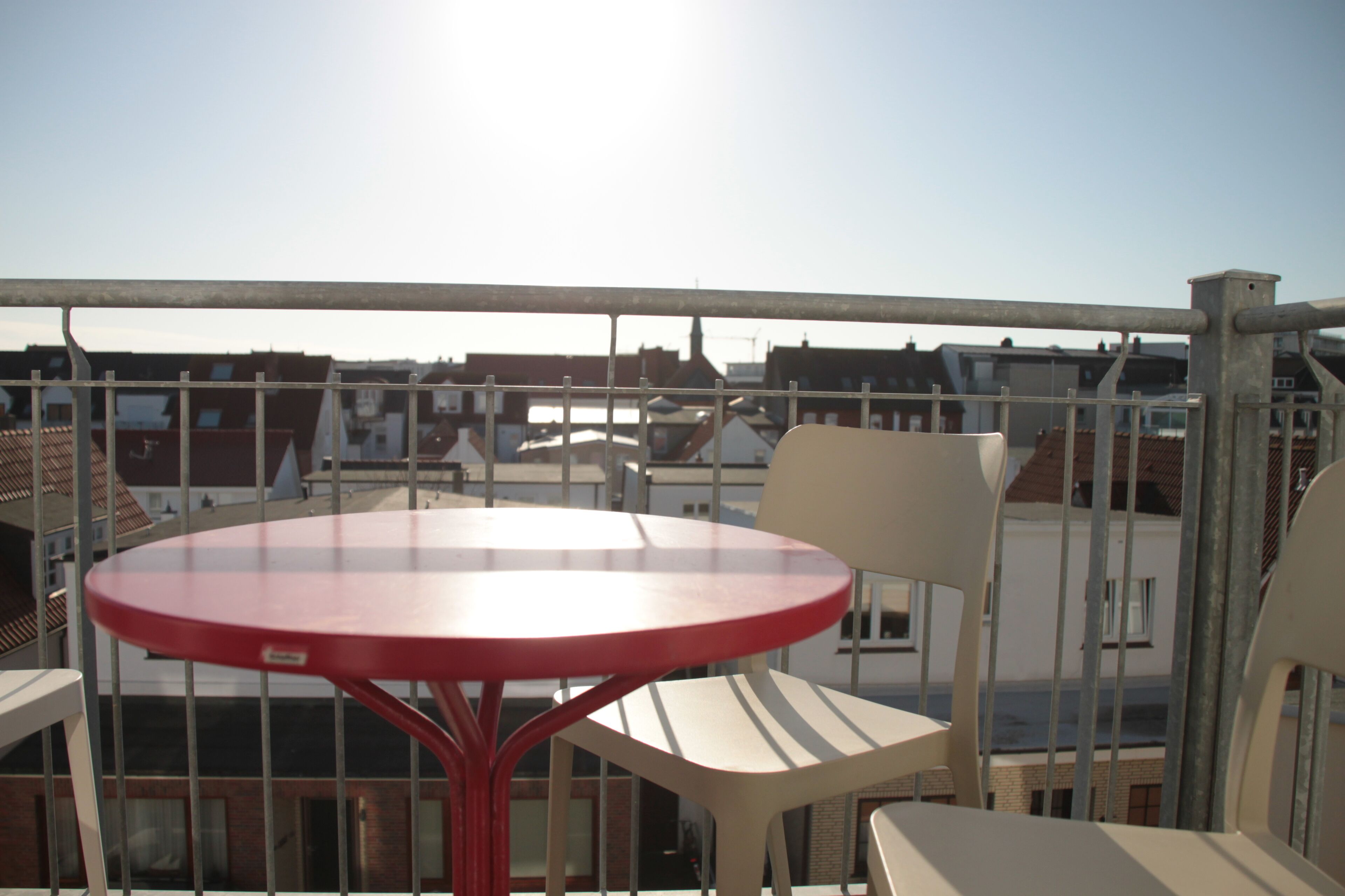 A red table and two chairs on a balcony with a view of buildings.