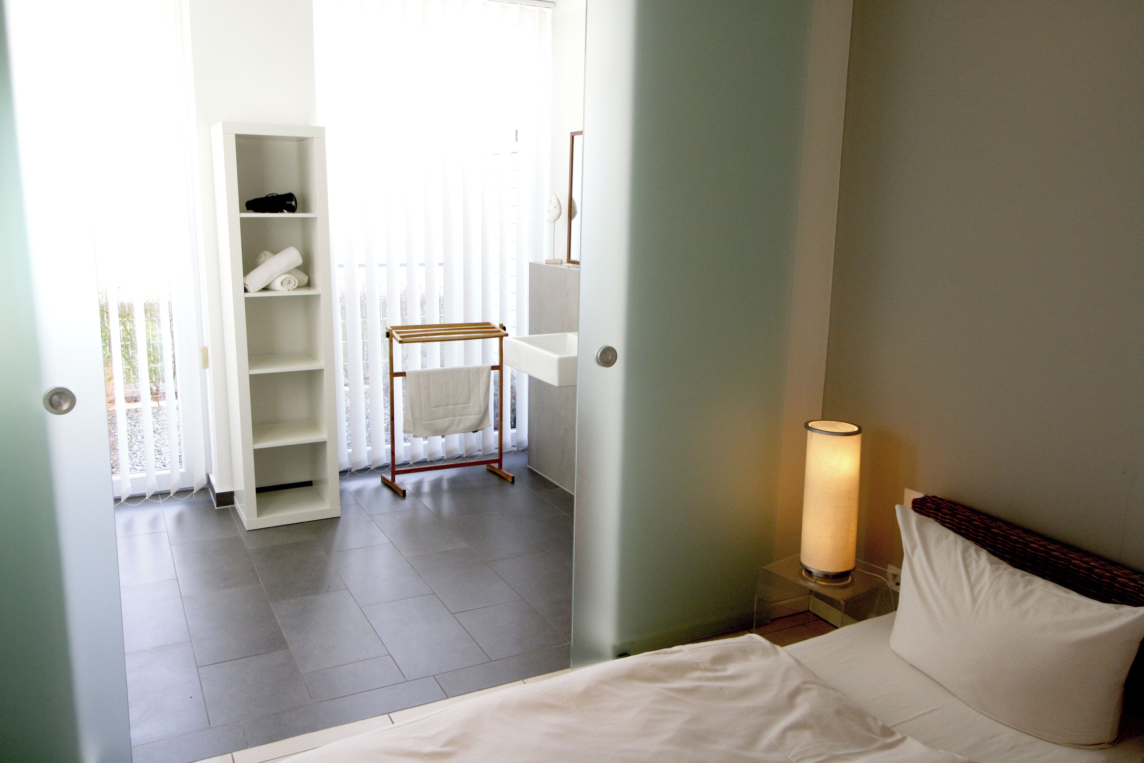 Habitación de hotel con cama, mesita de noche con lámpara, estante con toallas y vista al baño.