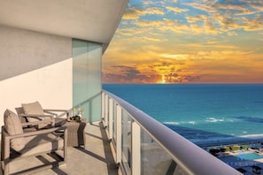 Un balcon avec vue sur l’océan au coucher du soleil, des meubles en osier et un espace piscine.