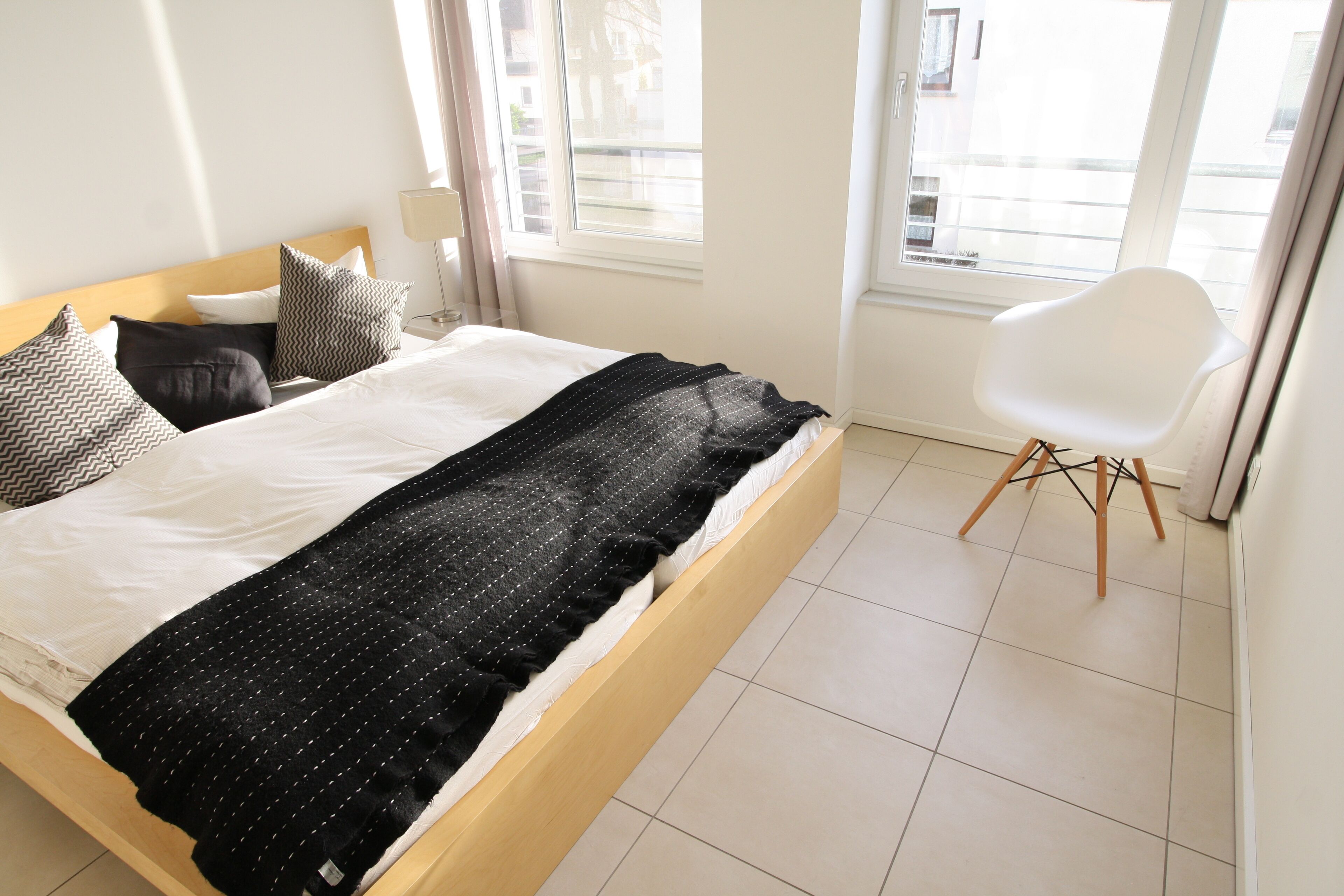 Un letto rifatto con cura, coperta a righe bianche e nere, una sedia bianca e una finestra con vista sugli edifici.