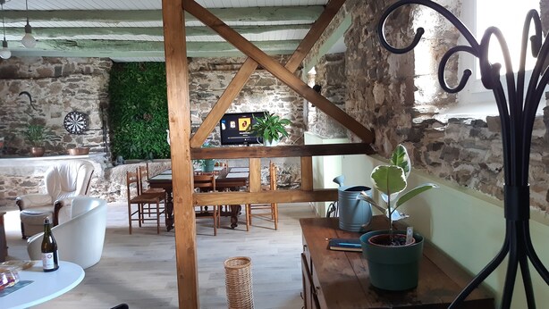 A rustic room with exposed stone walls, wooden beams, a dining area with chairs and a table, and a TV on a wooden stand. - L Abreuvoir DU Leffo Entre MER ET Campagne. Ressourcez Vous Avec vos Animaux (Allineuc)