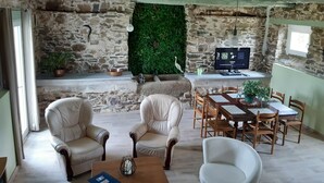 A rustic living room with stone walls, a green wall hanging, leather armchairs, a wooden dining table with chairs, and a television on a stand. - L Abreuvoir DU Leffo Entre MER ET Campagne. Ressourcez Vous Avec vos Animaux (Allineuc)