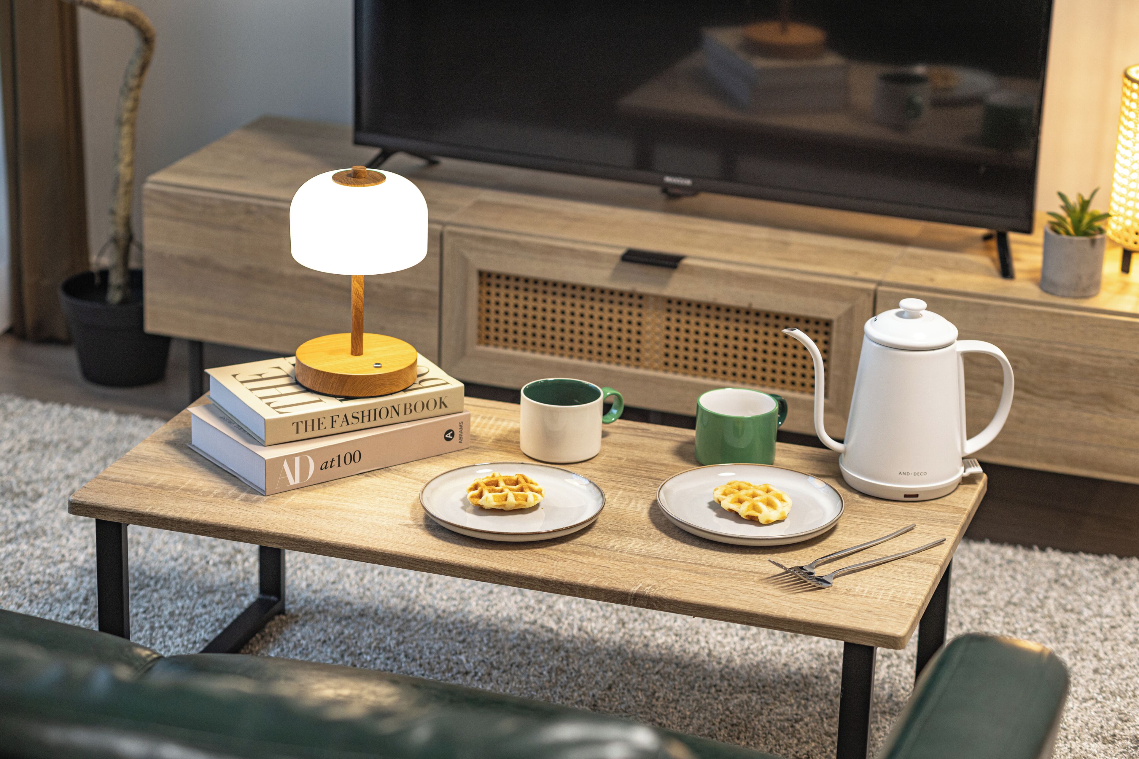 A small wooden table with a lamp, books, a mug, a plate with waffles, a teapot, and cutlery.