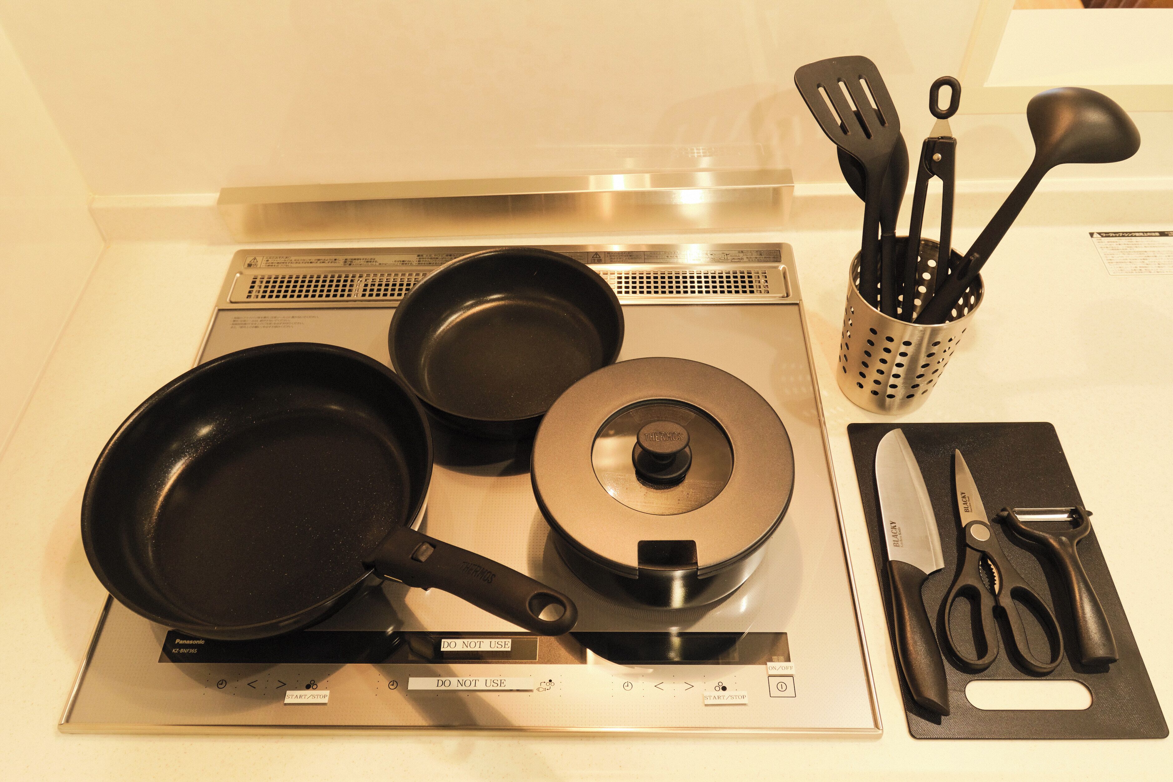 A kitchen with a stove, two frying pans, a pot, and a set of kitchen utensils including a knife, scissors, and a spatula.