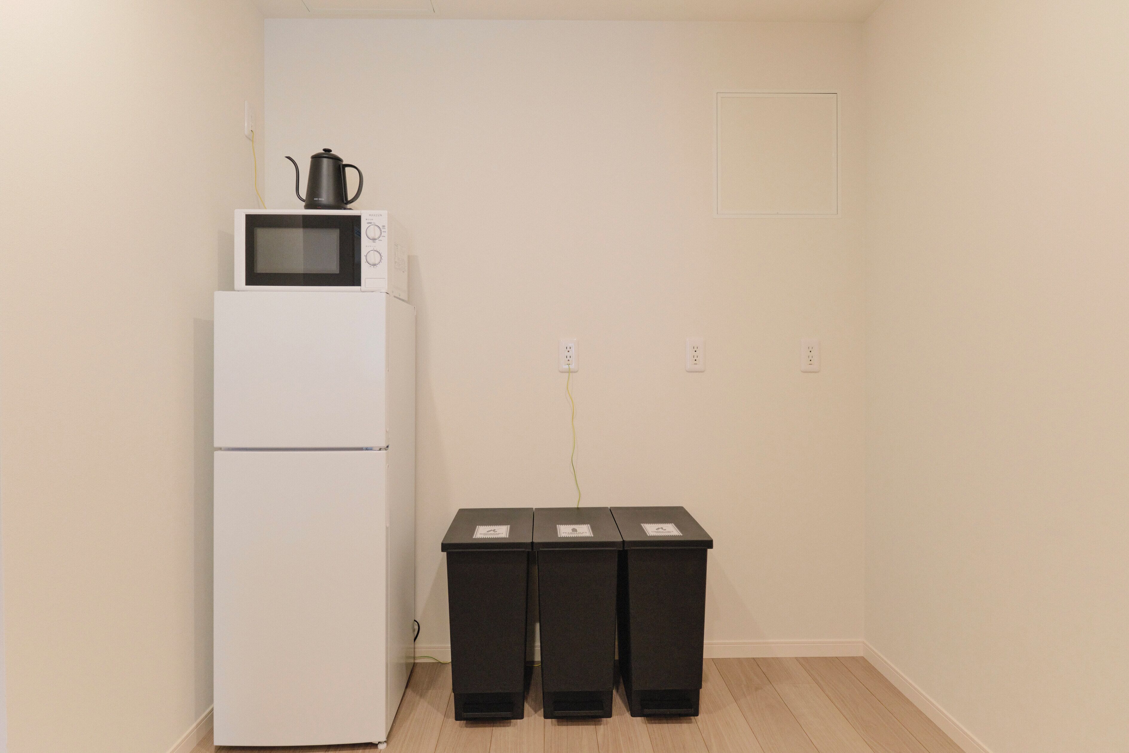 A kitchenette with a white refrigerator, a microwave, and a kettle on top. There are three black trash bins against the wall.