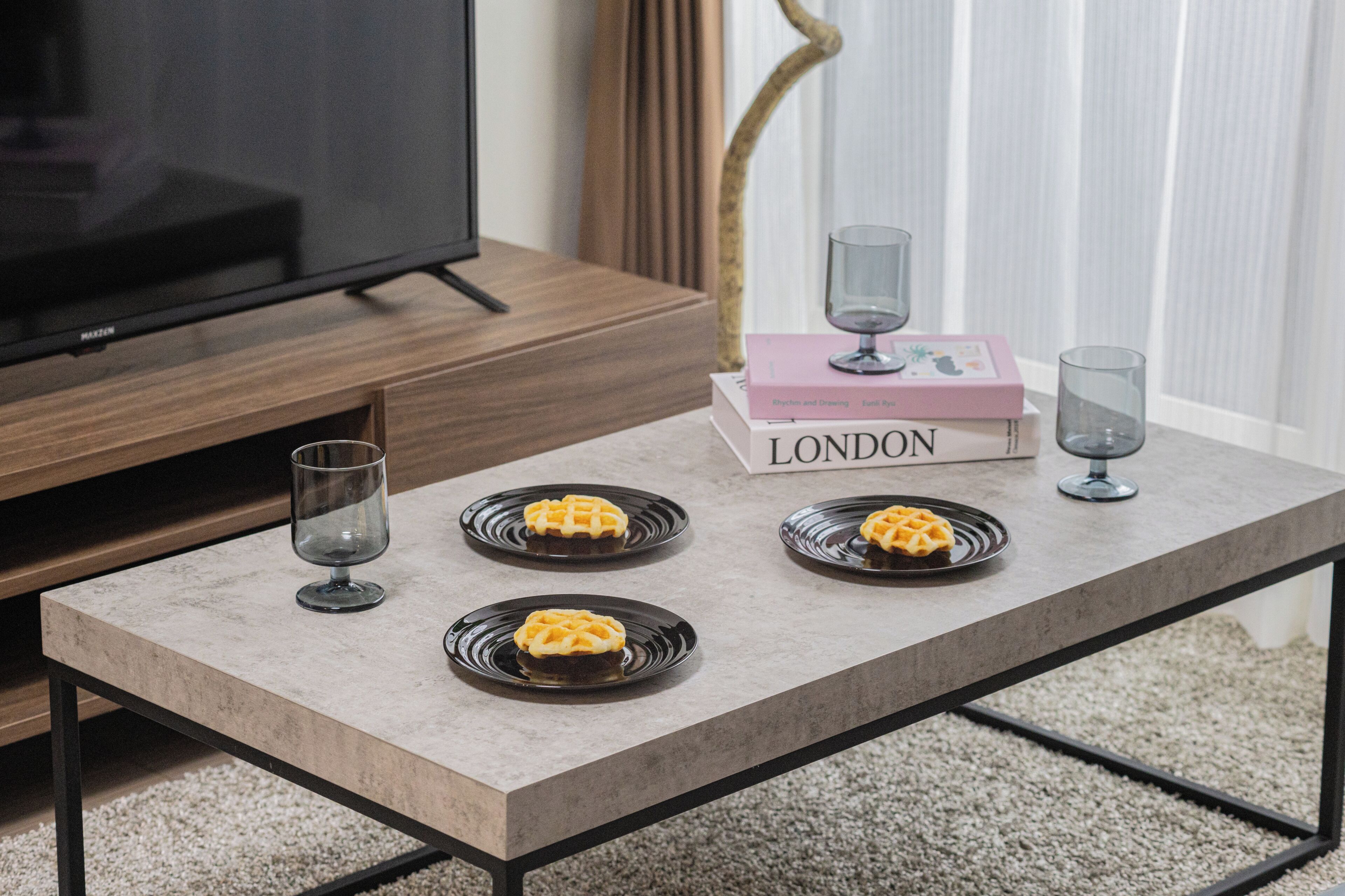 A modern living room with a flat-screen TV, a coffee table with two glasses and a book, and two plates with waffles.