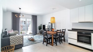 Ein modernes Wohnzimmer mit einer Couch, einem Esstisch mit Stühlen, einer Küchenzeile inklusive Schränken und Geräten sowie einem großen Fenster mit Vorhängen.