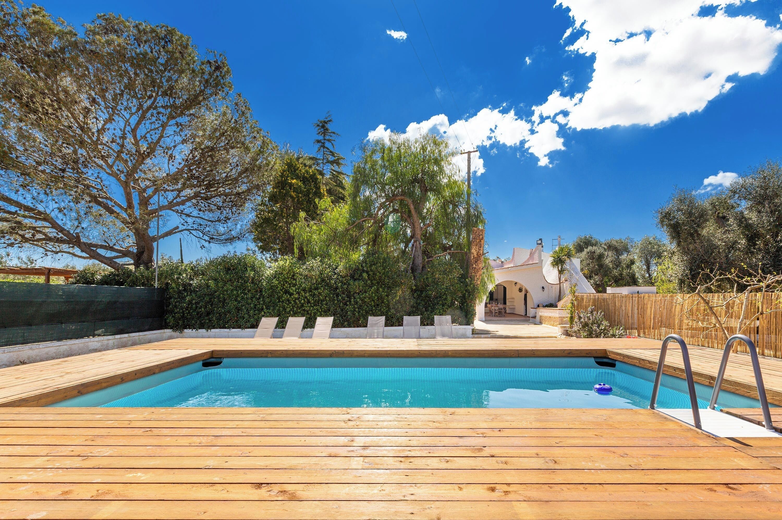 A wooden deck with a swimming pool, surrounded by trees and a clear blue sky.