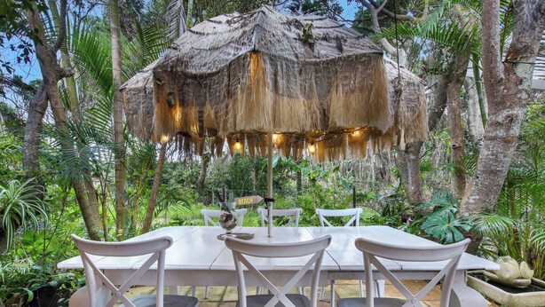A dining area with a thatched roof, white chairs, and a table set outdoors surrounded by lush greenery. - Shellies on the Beach Little House - 2BR Absolute Beachfront with Outdoor Bath (Sawtell)