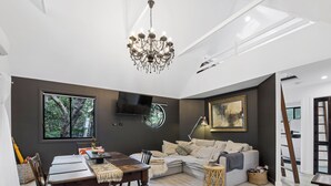 A modern living room with a dining area, a chandelier, a flat-screen TV, and a large window with a view of trees. - Shellies on the Beach Little House - 2BR Absolute Beachfront with Outdoor Bath (Sawtell)