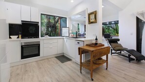 A modern kitchen with white cabinets, a built-in oven, and a large window. There is a wooden table with a chair and a small sofa in the living area. - Shellies on the Beach Little House - 2BR Absolute Beachfront with Outdoor Bath (Sawtell)