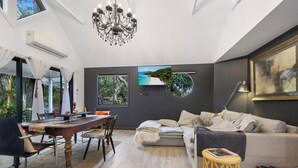 A modern living room with a dining area, a chandelier, a flat-screen TV, and a large window with a view of the beach. - Shellies on the Beach Little House - 2BR Absolute Beachfront with Outdoor Bath (Sawtell)
