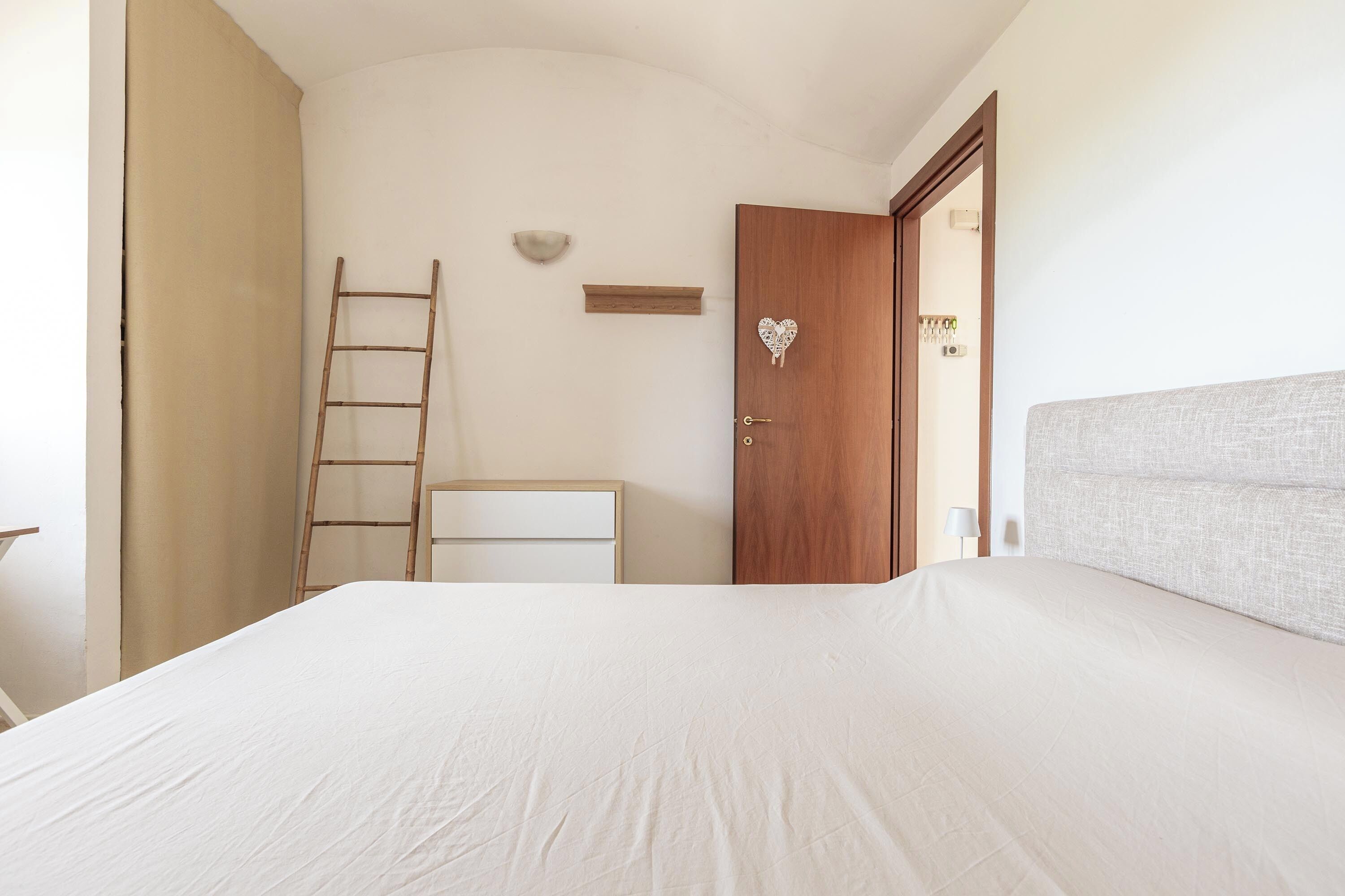 A bedroom with a bed, a wooden door, a ladder, a bedside table, and a wall-mounted light fixture.