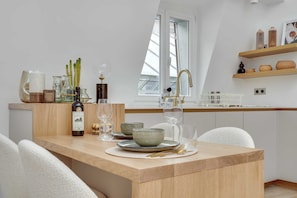 A modern kitchen with a wooden dining table set for two, a window with a view, and a shelf with kitchenware. - Résidence Paris CHOISEUL (Paris)