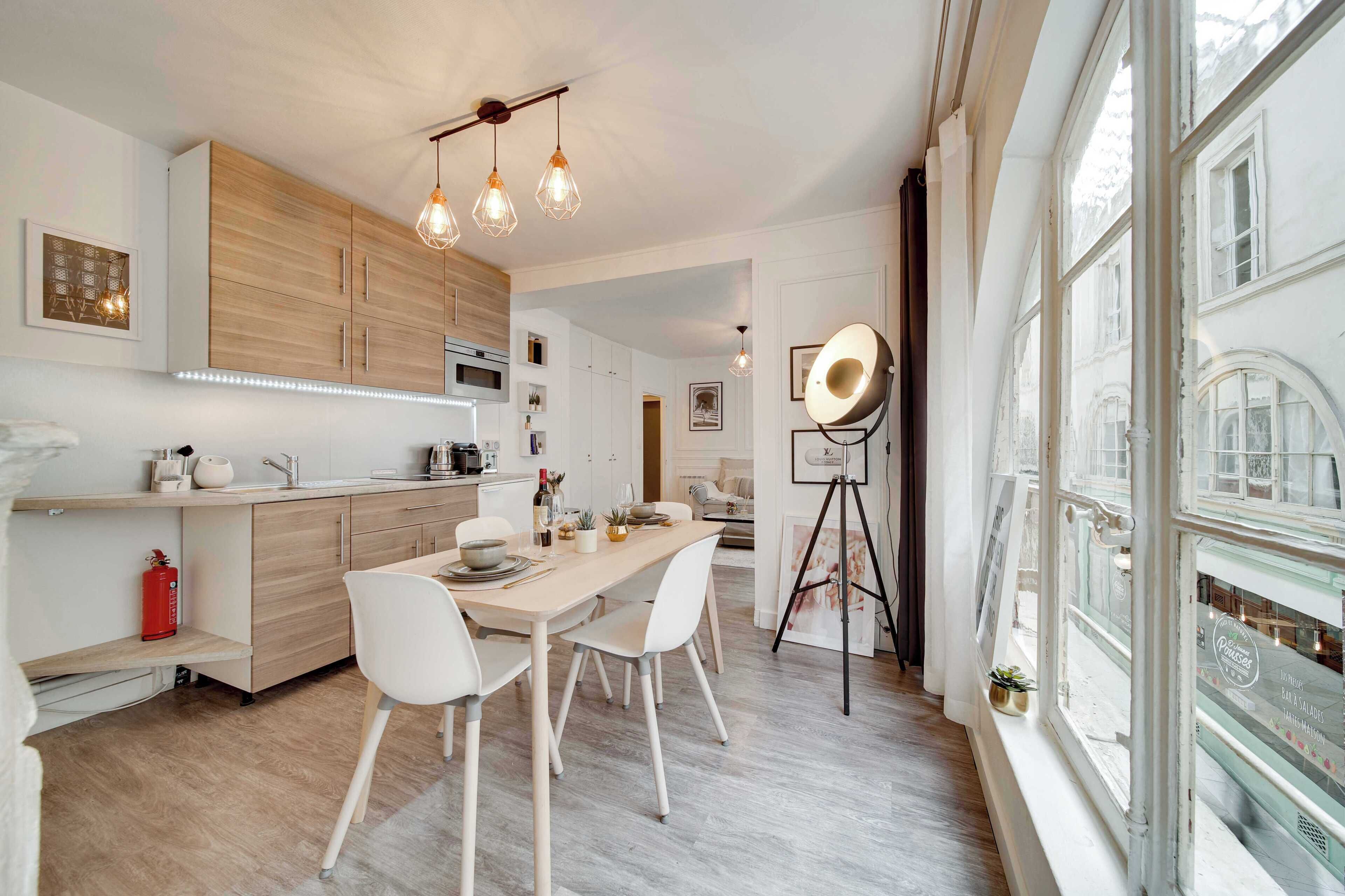 Una cocina moderna con armarios de madera, una mesa de comedor con sillas, iluminación colgante y un ventanal con vistas a edificios.