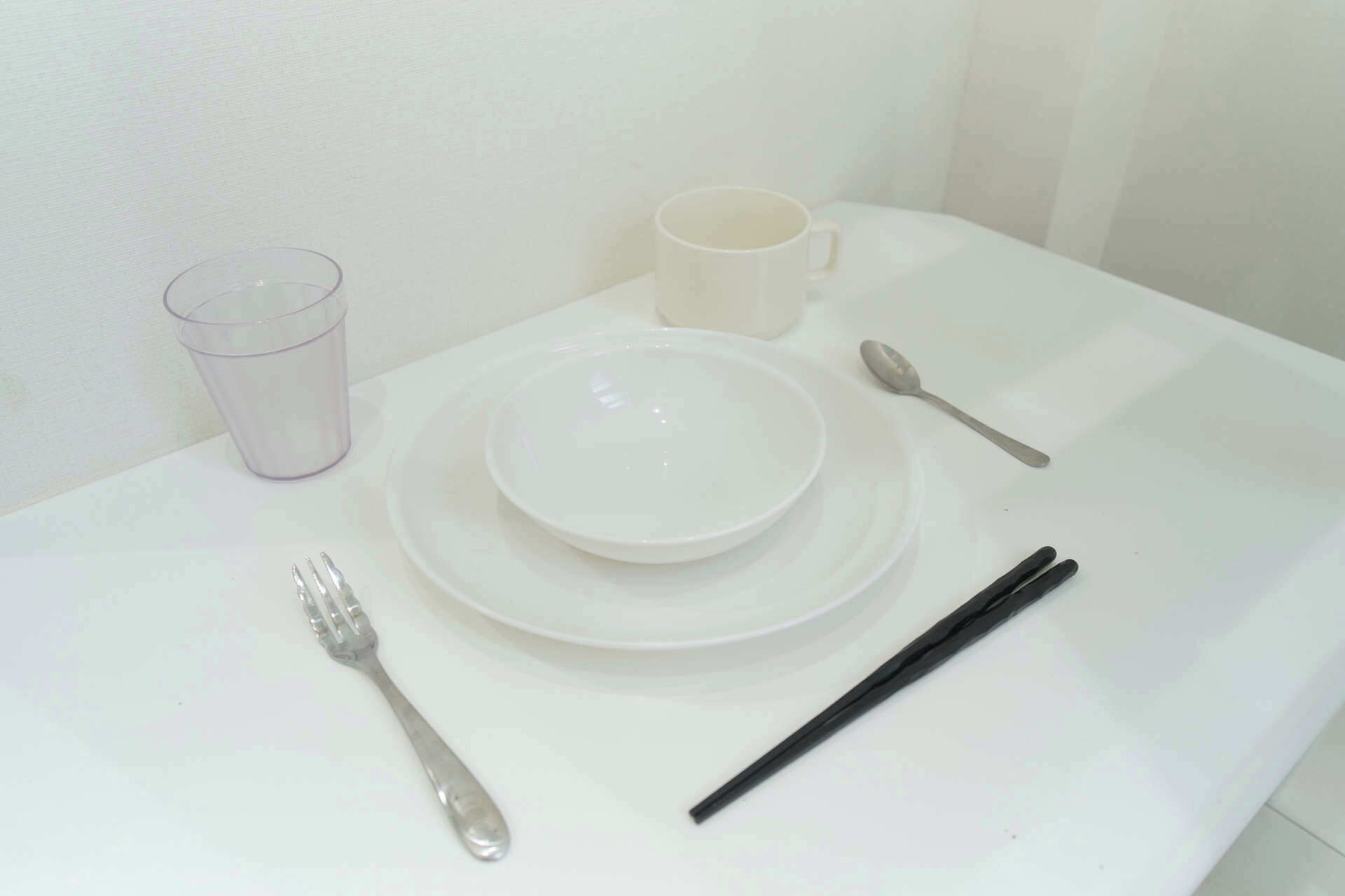 A white table with a white plate, a white cup, a fork, a spoon, and chopsticks.
