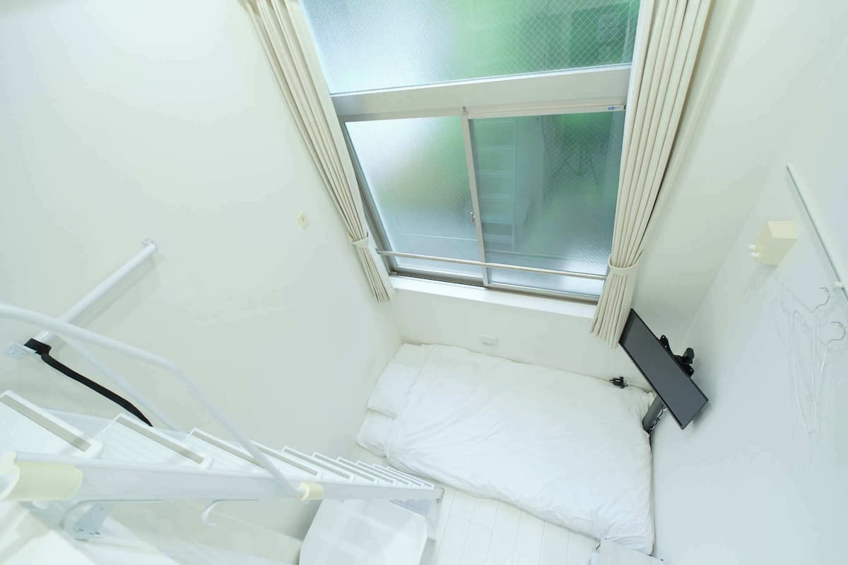 A small, narrow room with a single bed, a window with curtains, and a wall-mounted TV.