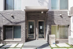 A modern building entrance with a glass door, a small brick pillar with a sign, and a window with a metal grill. - Nano Kamiochiai (Tokyo)