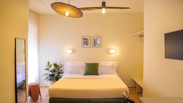 Quarto de hotel moderno com uma cama grande, mesas de cabeceira, um banquinho pequeno, uma planta em vaso e uma televisão de tela plana.