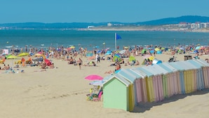 Ein überfüllter Strand mit Menschen, die sich sonnen und schwimmen. Bunte Sonnenschirme und Strandkörbe sind zu sehen.
