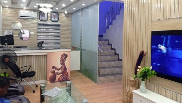 A modern reception area with a desk, computer, and framed pictures. A staircase with a blue-lit landing is visible in the background. - Tristar Hotel & Suites  (Lekki)