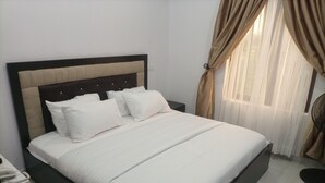 A neatly made bed with white bedding and a dark headboard, a bedside telephone, and a window with curtains. - Tristar Hotel & Suites  (Lekki)