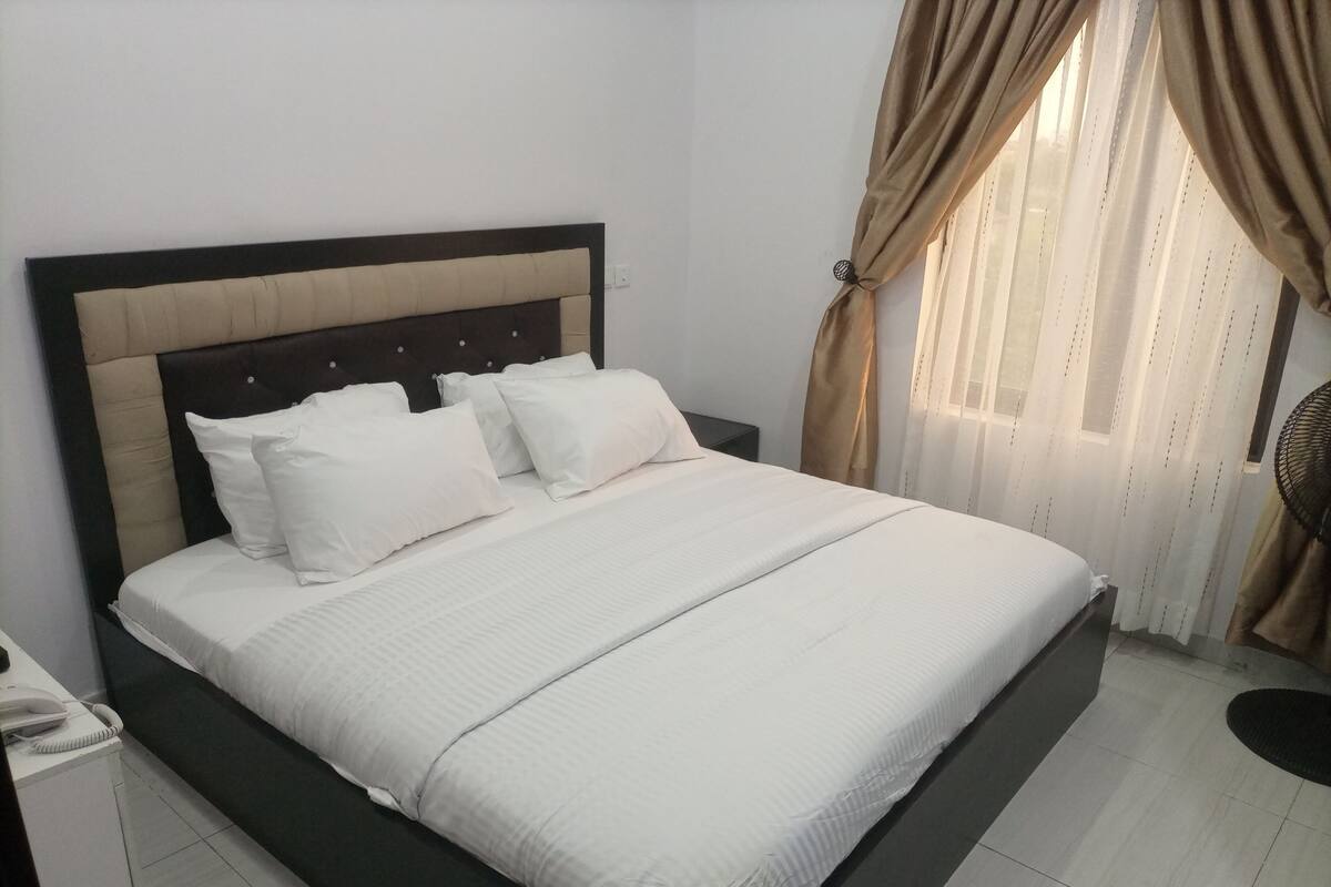 A neatly made bed with white bedding and a dark headboard, a bedside telephone, and a window with curtains.