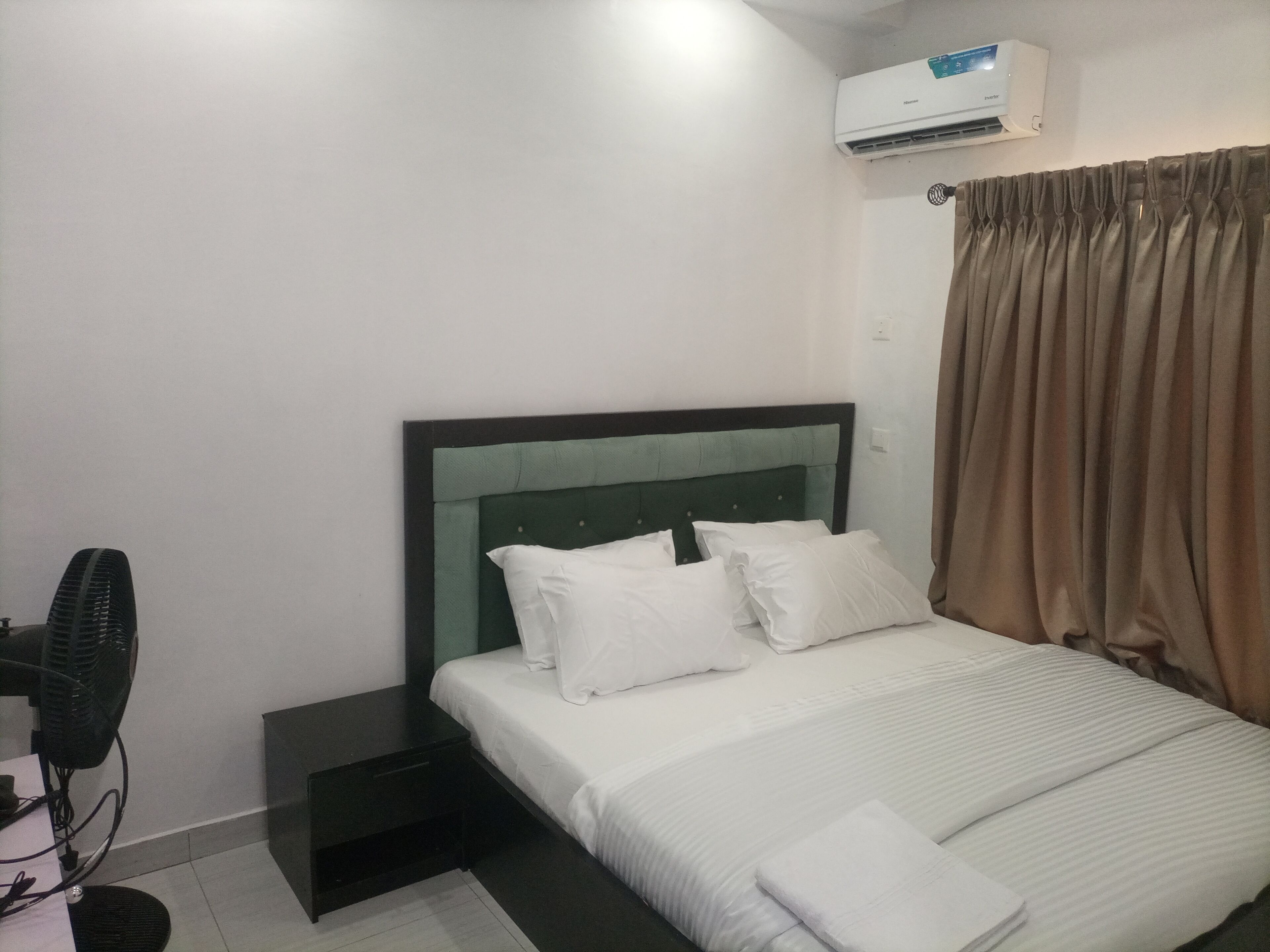 A hotel room with a bed, a bedside table, an air conditioning unit, a fan, and a curtain.
