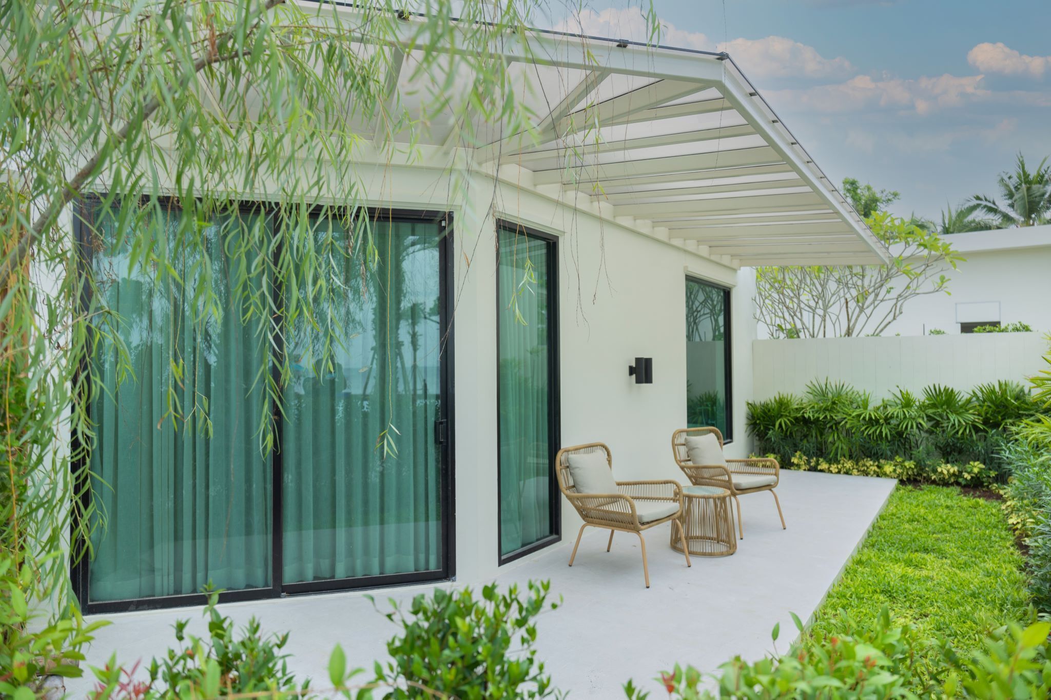 A modern house with large sliding glass doors, a covered patio with wicker furniture, and a well-maintained garden.