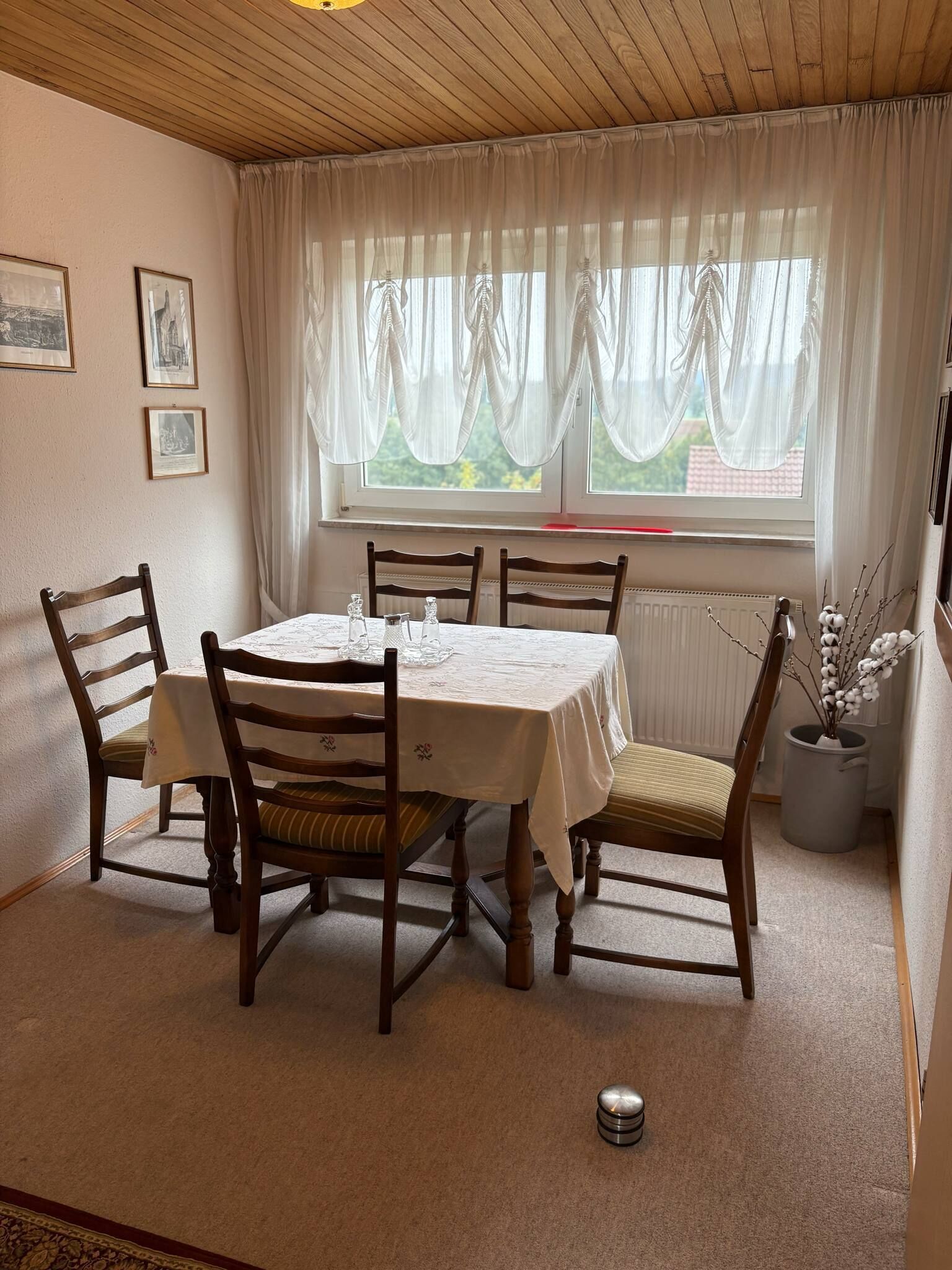 Un coin repas avec une table ronde, quatre chaises en bois, une nappe blanche et une fenêtre ornée de rideaux légers.