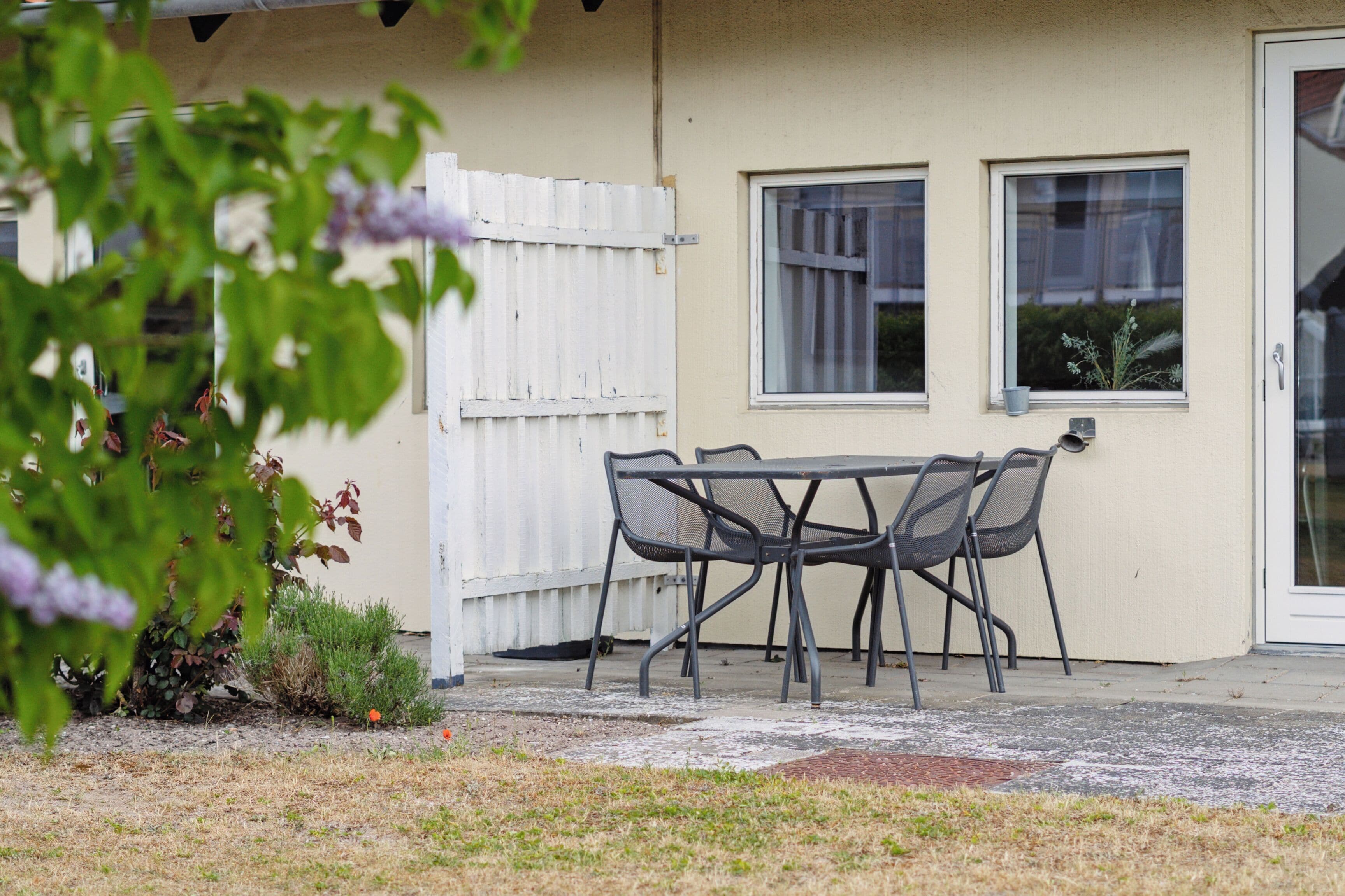 A patio with a table and chairs, a white fence, and a building with two windows.