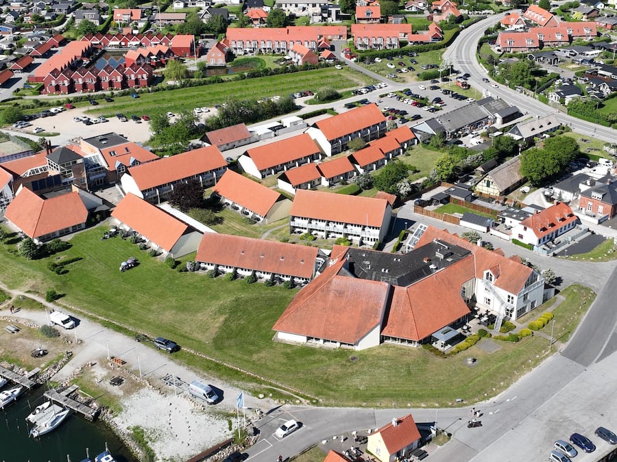An aerial view of a residential area with red-roofed houses, a marina with boats, and a road curving through the neighborhood.