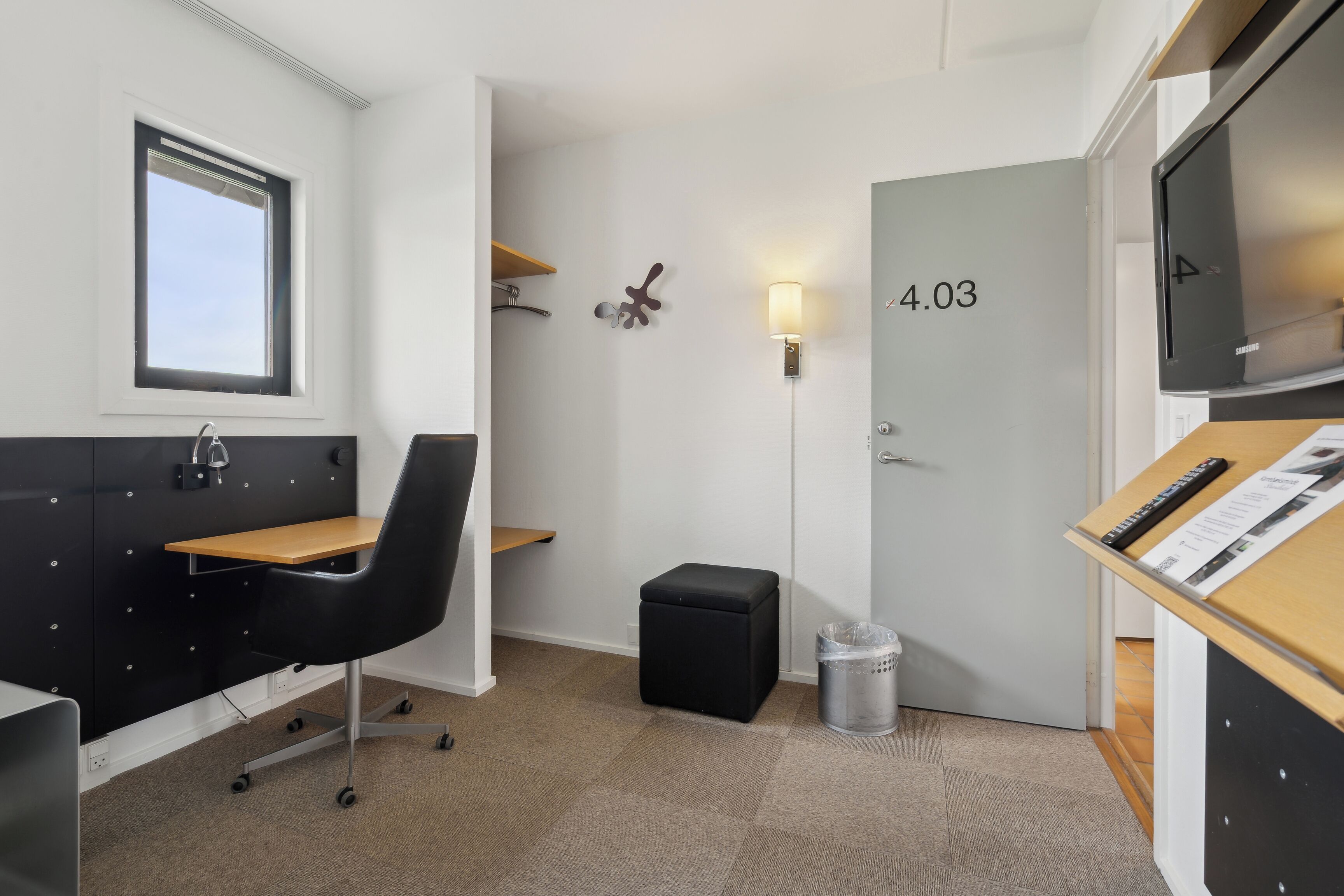 Ett kompakt hotellrum med ett skrivbord, en stol och en liten pall. En papperskorg står placerad nära dörren, och en vägglampa syns.