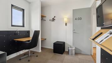 Ett kompakt hotellrum med ett skrivbord, en stol och en liten pall. En papperskorg står placerad nära dörren, och en vägglampa syns.