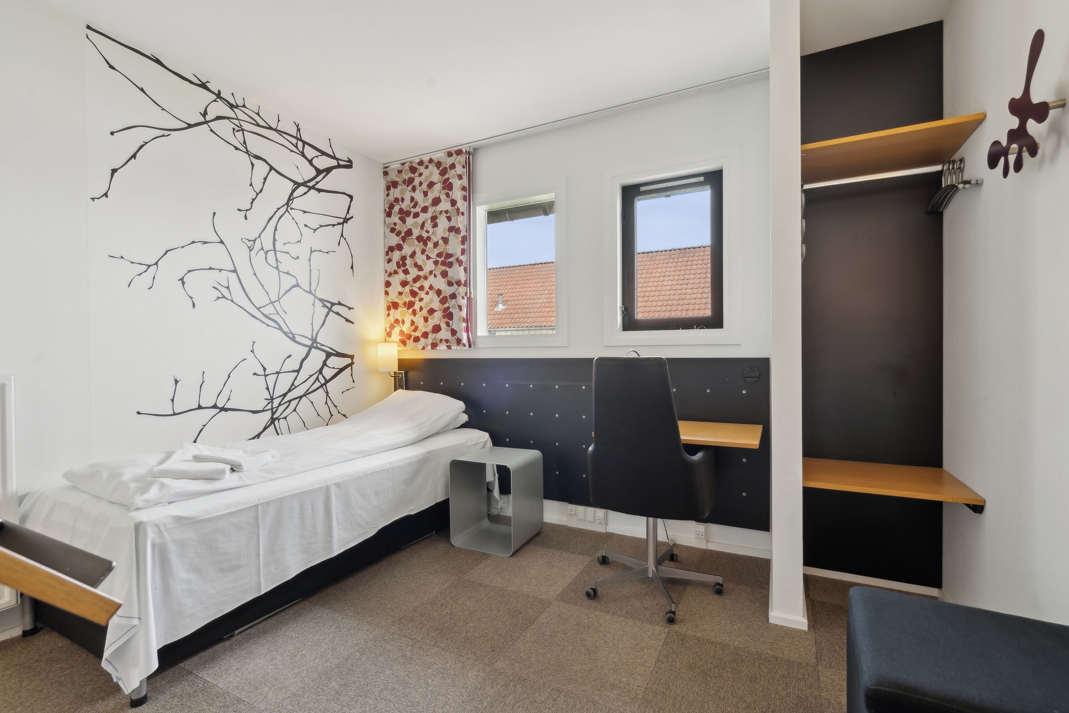 Een kleine kamer met een bed, een bureau en een stoel. Er zijn twee ramen en een wandplank.